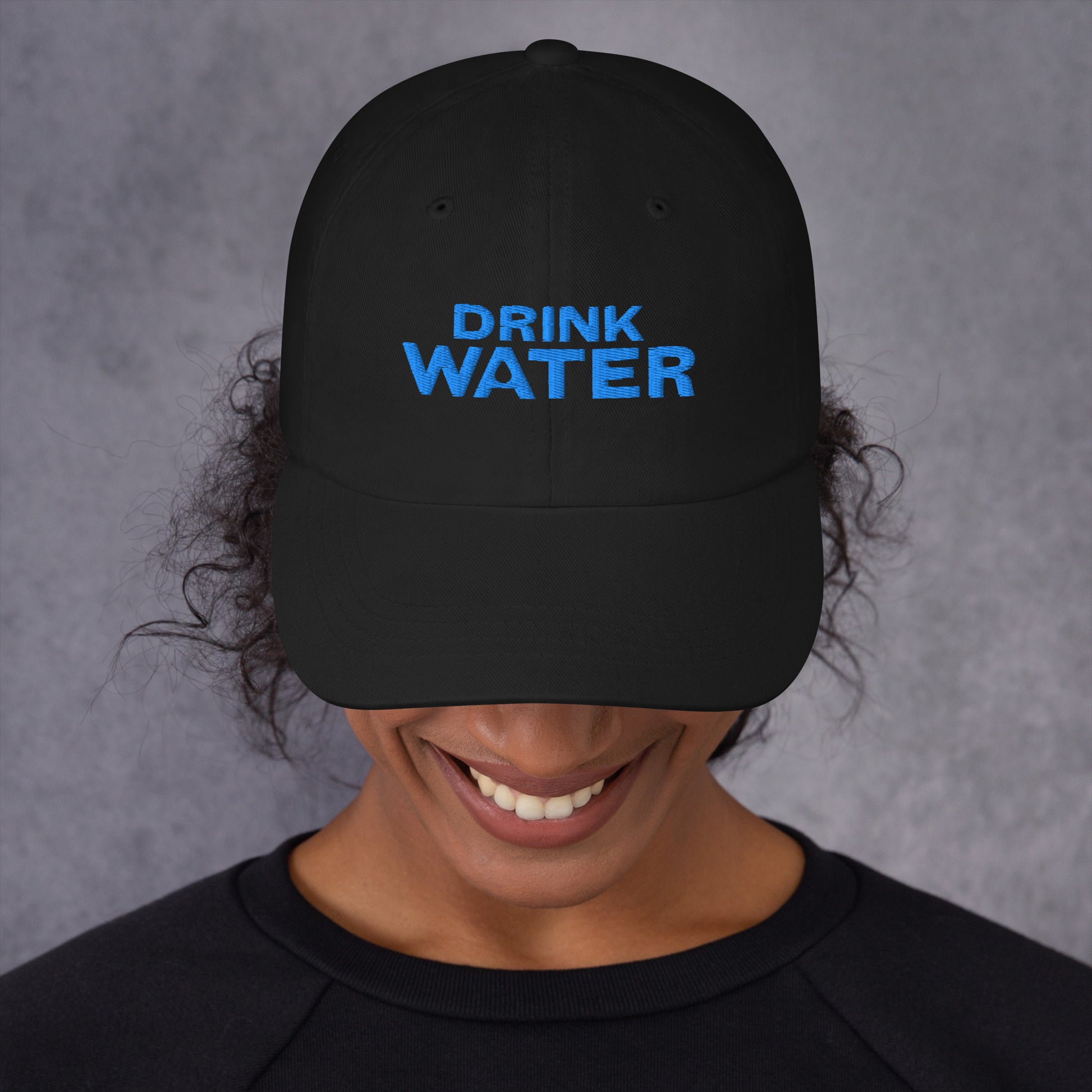 Black classic dad hat with bright blue text that reads ‘DRINK WATER’ designed by queer creators to express the need to remember to stay hydrated. Front view, on model.
