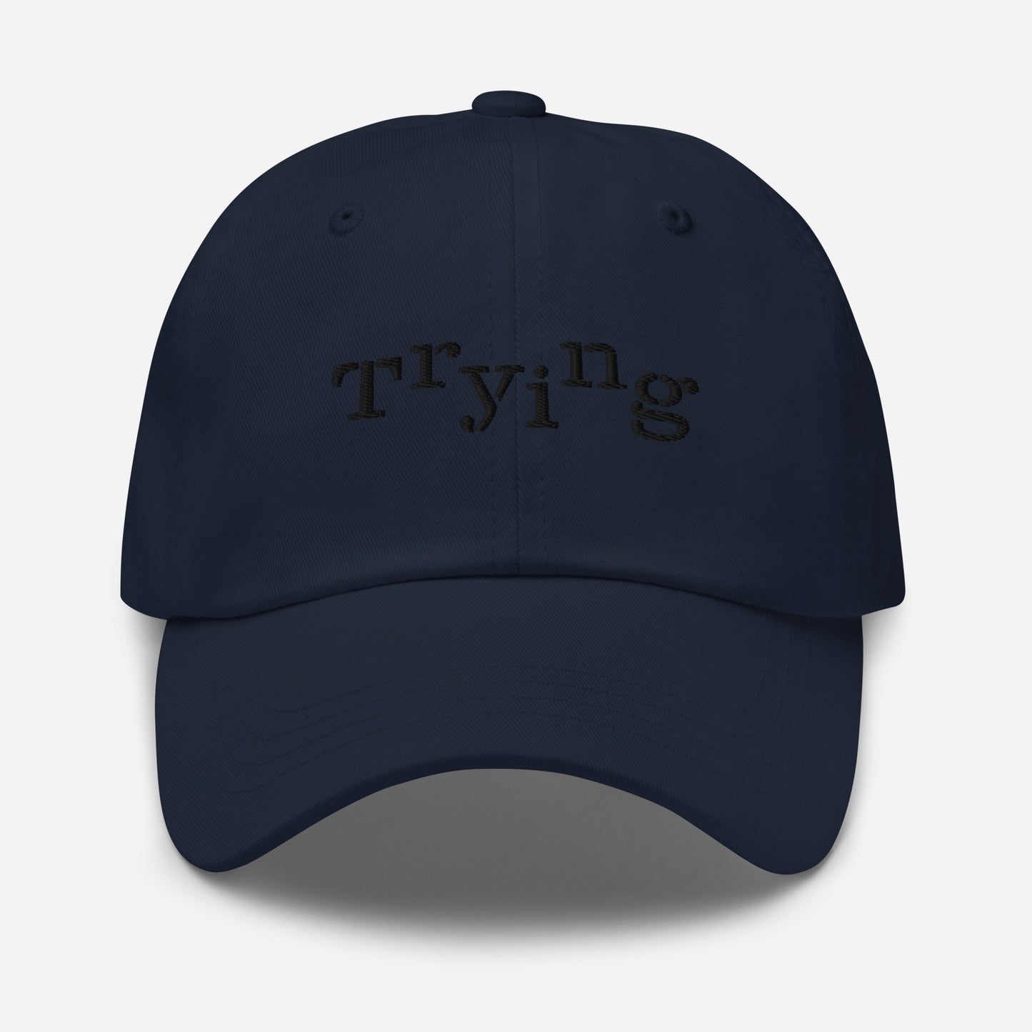 Navy classic dad hat with black embroidered text that reads ‘Trying,’ designed by queer creators to express mental health and perseverance. Front view, not on model.