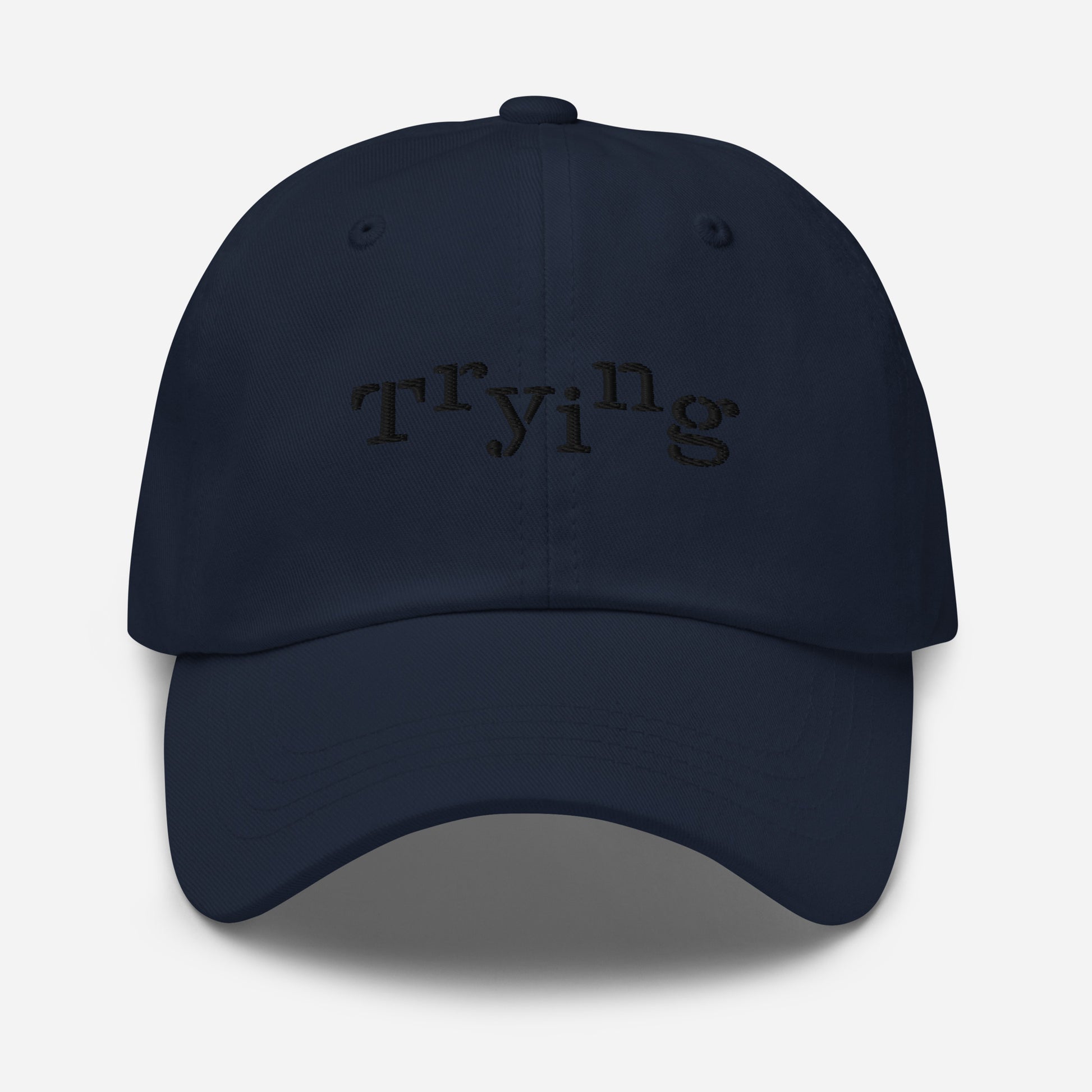 Navy classic dad hat with black embroidered text that reads ‘Trying,’ designed by queer creators to express mental health and perseverance. Front view, not on model.