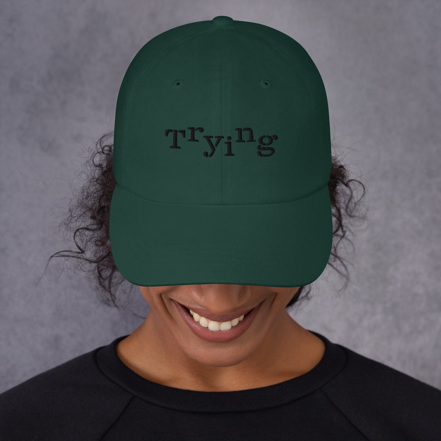 Spruce classic dad hat with black embroidered text that reads ‘Trying,’ designed by queer creators to express mental health and perseverance. Front view, on model.