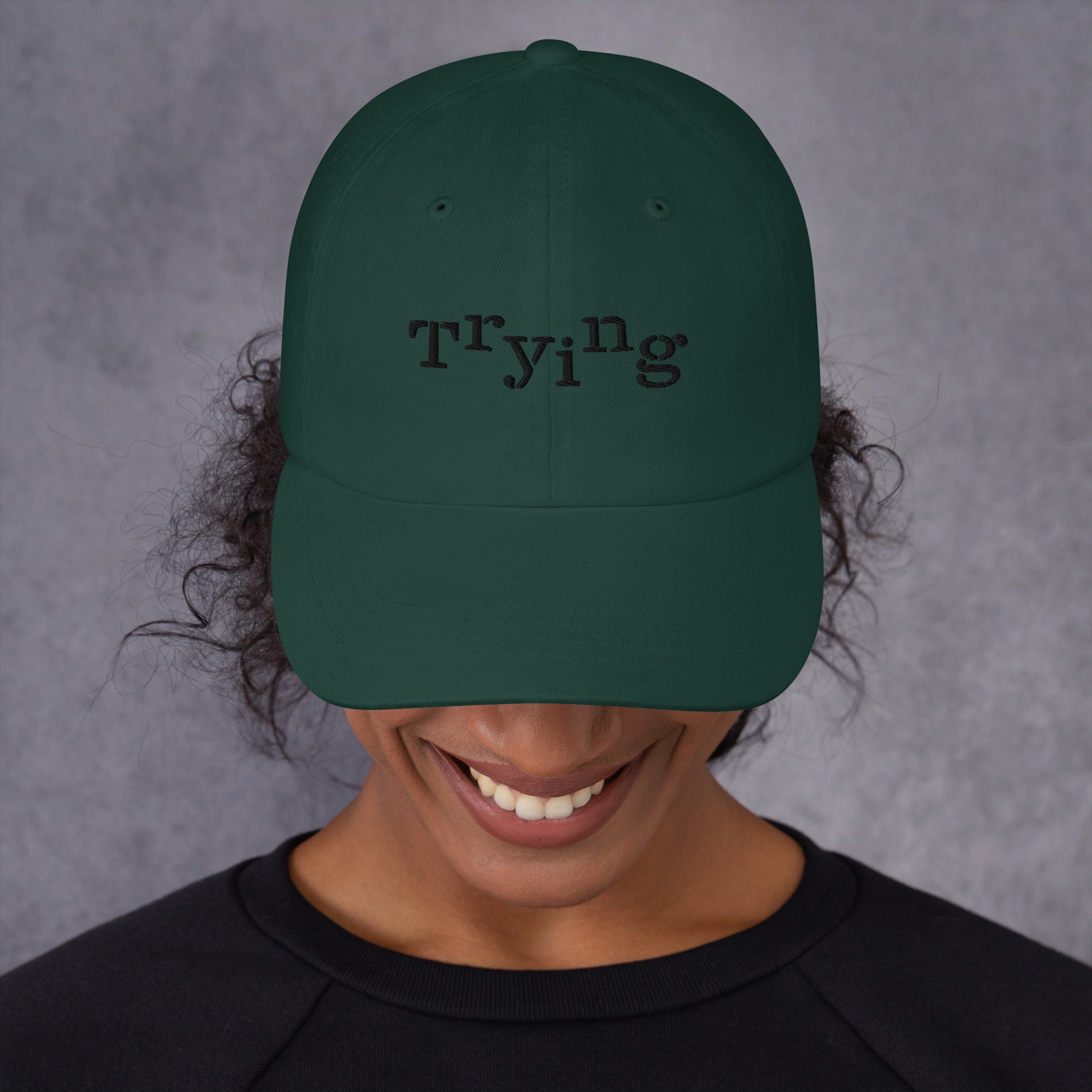 Spruce classic dad hat with black embroidered text that reads ‘Trying,’ designed by queer creators to express mental health and perseverance. Front view, on model.