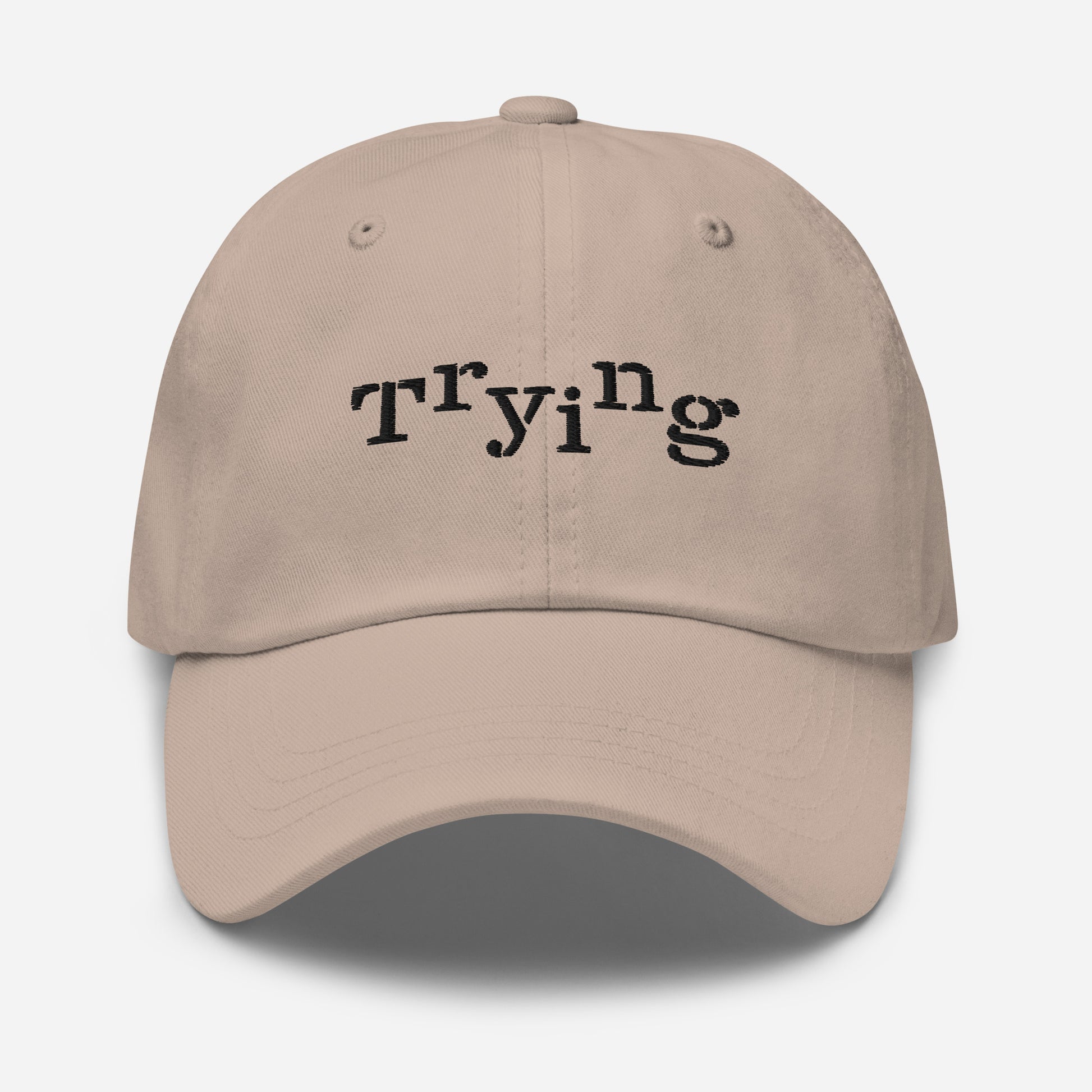 Stone classic dad hat with black embroidered text that reads ‘Trying,’ designed by queer creators to express mental health and perseverance. Front view, not on model.